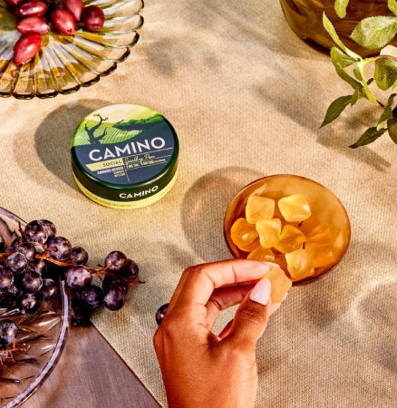 Camino Sparkling Pear gummies in a green container with a hand reaching for a gummy, surrounded by grapes and foliage.