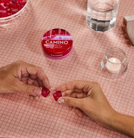 Hands holding Camino Wild Cherry gummies with the product container on a pink tablecloth, featuring a candle and glass of water.