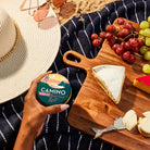 Hand holding Camino Watermelon Lemonade Bliss gummies next to a cheese board with grapes and a sun hat on a beach blanket.
