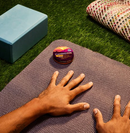 Small round tin of Camino Sours hemp-infused sour candies on yoga mat, with blue box and colorful blanket nearby.
