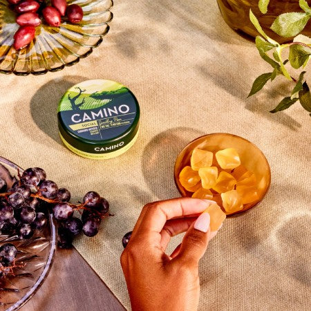 Camino Sparkling Pear gummies in a green container with a hand reaching for a gummy, surrounded by grapes and foliage.