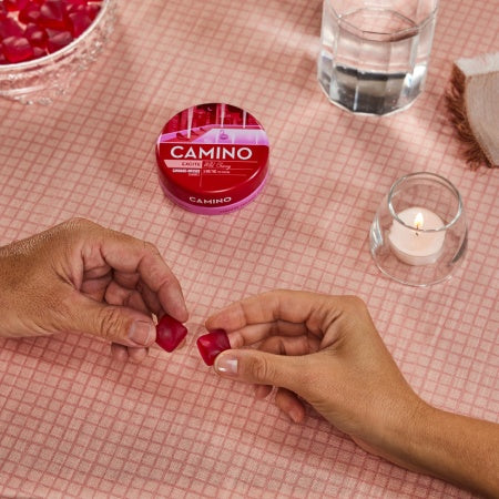 Hands holding Camino Wild Cherry gummies with the product container on a pink tablecloth, featuring a candle and glass of water.