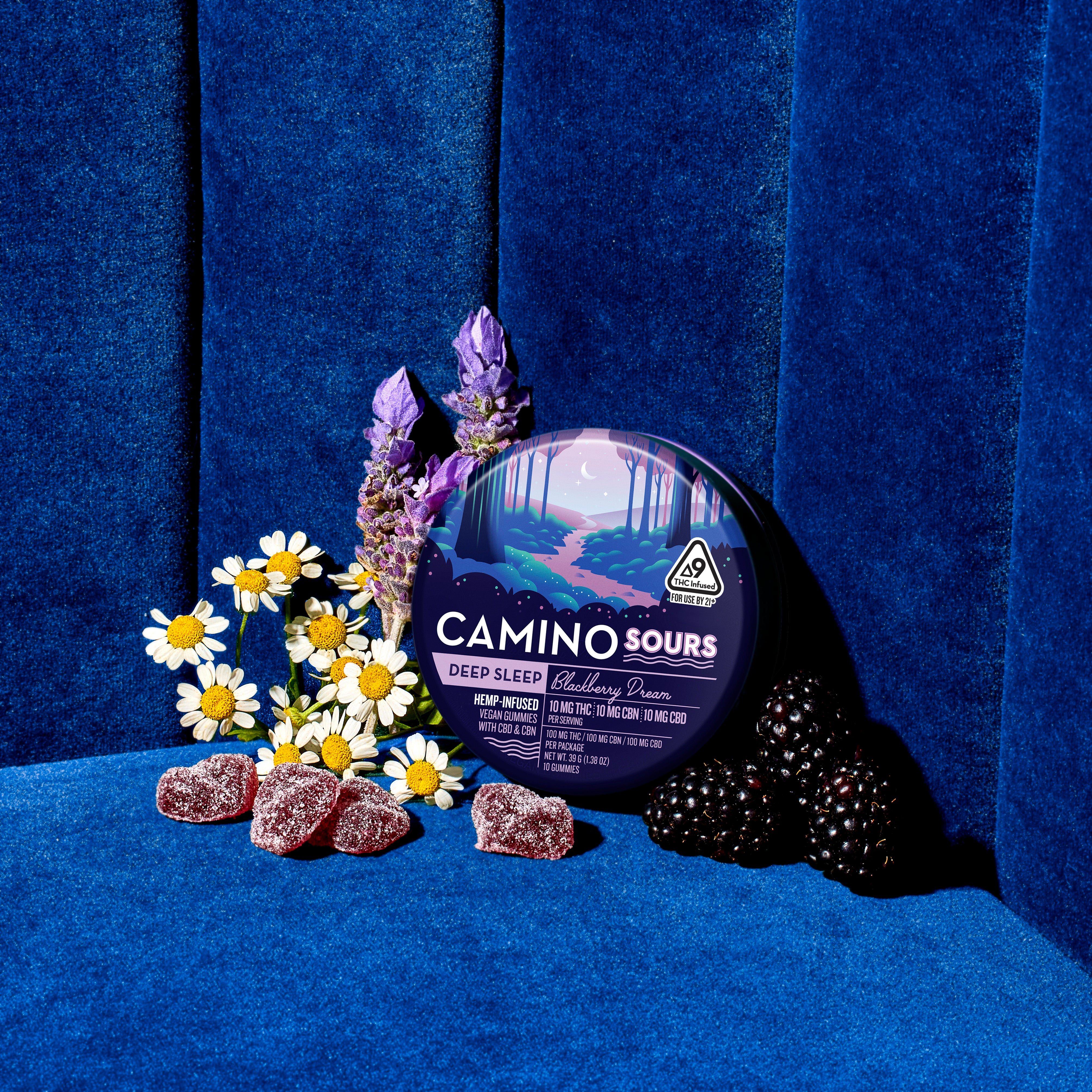 Blue container of Camino Sours Deep Sleep gummies with blackberry flavor, surrounded by flowers and blackberries, on a blu...