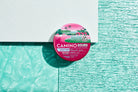 Pink and green watermelon spritz flavored hemp-infused gummies in a round container with tropical ocean and palm tree desi...